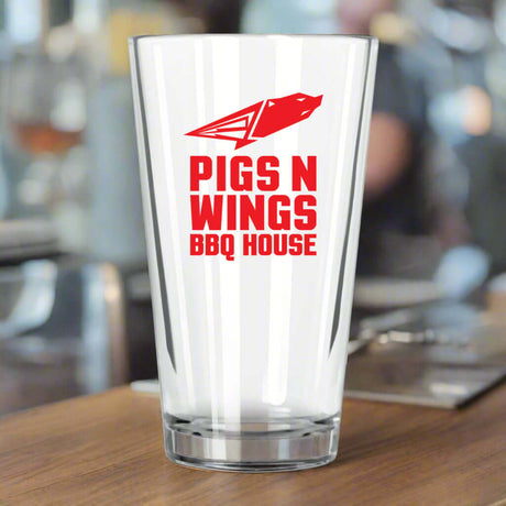 Custom Libbey 20 oz pint glass featuring 'Pigs N Wings BBQ House' logo in red on a wooden table.