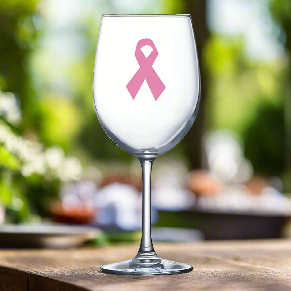 Custom wine glass with pink ribbon design set in a natural outdoor background.